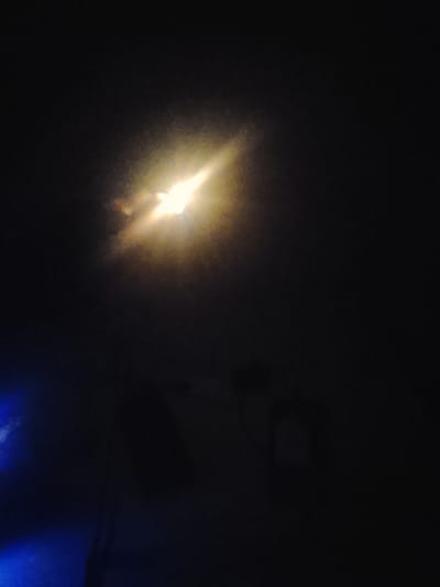 This is a photograph taken at night in Orodo, Nigeria. The image is predominantly dark, with a bright, yellowish light source dominating the upper center of the frame. This light appears to be a powerful artificial light, possibly a spotlight or a strong flashlight, casting a glow and illuminating a hazy atmosphere.

In the lower left quadrant, there is a distinct area of blue light, suggesting another light source or a reflection. The background is very dark and largely indistinct, hinting at a nighttime scene, likely outdoors due to the nature of the light. There are faint, blurry shapes in the background that could be structures or objects, but they are not clearly discernible.

The overall mood is somewhat mysterious and dramatic, due to the contrast between the intense light and the deep darkness. There are no visible people or animals. The scene appears to be set in a location where such lighting might be used for illumination or signaling. The time of day is definitively night. There are no weather cues, but the haziness around the light source might suggest some atmospheric conditions like mist or smoke. No text is visible in the image.