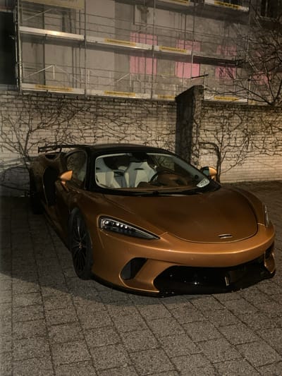 A metallic bronze McLaren GT sports car with a black roof and white interior is parked at night on a paved surface in Dietlikon, Switzerland. The vehicle features black multi-spoke wheels, visible LED headlights, and a rear spoiler. Behind the car is a light-colored brick wall covered with leafless vines. Further in the background, a building under construction or renovation is obscured by metal scaffolding, displaying yellow banners with the text "huberbeugeschgeschafft.ch" and "Telefon 044 211 50 80." Pink insulation or covering material is visible on parts of the building through the scaffolding. No individuals, actions, or interactions are present in the scene.