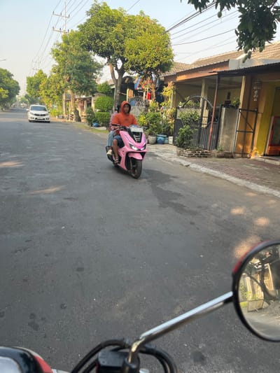 A person wearing an orange hooded sweatshirt and blue jeans rides a pink scooter down a paved street in a residential area. The scooter has a license plate that reads "W 2451 NHP". The person is looking forward, and the scooter is moving away from the camera. In the background, there are houses with green trees and manicured bushes. A white car is parked on the side of the road to the left of the scooter. The sky is clear and bright. This scene appears to be located in Karangtanjung, Indonesia, as indicated by the license plate format and the typical residential architecture and vegetation seen in the image.