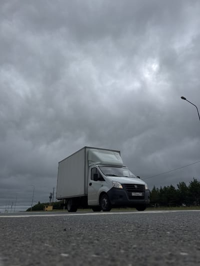 A white box truck is driving on a two-lane asphalt road under a cloudy sky. The truck is in the right lane, and its headlights are on. The road has white lane markings. On the left side of the road, there are streetlights and a line of trees. In the distance, there is a small yellow structure. The license plate of the truck is visible and reads "T134AH 716". The image appears to have been taken in the city of Alnashi, Russia.
