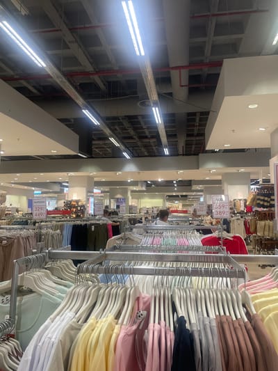 The image, taken in Bağcılar, Turkey, depicts the interior of a brightly lit retail store, likely a clothing boutique or department store. The perspective is tilted, offering a view of clothing racks filled with a variety of garments, predominantly tops and sweaters in pastel colors like pink, yellow, light blue, and beige, alongside darker options like navy and brown. The clothing is neatly arranged on hangers, suggesting an organized retail environment.
In the background, rows of shelving units are visible, some of which appear to be empty. Industrial-style lighting fixtures are prominently featured along the ceiling, casting a strong illumination. Red pipes and conduits are also visible, integrated into the ceiling infrastructure.
There are a few individuals present, though their features are not clearly discernible. One person, likely a customer, is browsing the clothing racks, partially obscured by the garments. Another person appears to be standing further back in the aisle.
Several price tags are visible, displaying prices in Turkish Lira (TL). One tag reads "359,99 TL" and another "399,99 TL". Another tag has "329,99 TL" and "449,99 TL". Some tags also include text such as "BODY" and "PANTOLON".
The overall impression is of a typical commercial setting, with an emphasis on the display of merchandise. The time of day and weather conditions are not discernible from the image.