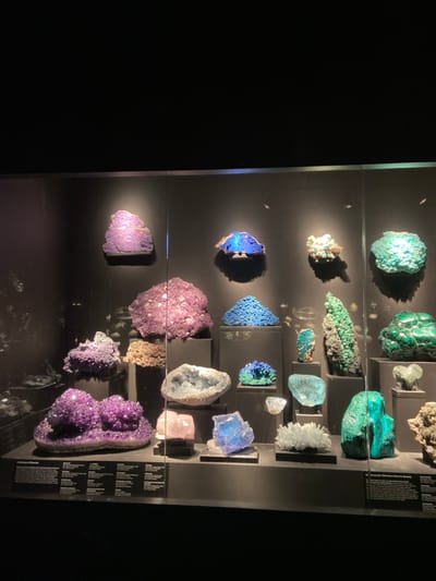 The image shows a glass display case containing 15 mineral specimens in a museum at the American Museum of Natural History located in New York City at the coordinates 41.02, -73.623. The display case is lit from above and contains several different types of minerals, including amethyst, malachite, azurite, and fluorite. The specimens are arranged in a random pattern, and are separated by black velvet-lined shelves. The glass case has a dark background, which makes the minerals stand out. The minerals are all brightly colored and have a variety of shapes and sizes. Some of the minerals have been polished, while others have not. The image was taken with a phone camera, and the image is of medium quality. The scene is lit by artificial light. There are no people or other animals in the image.