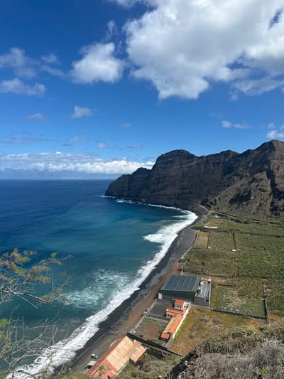 This image depicts an elevated view of a coastal landscape in Hermigua, Spain. The upper portion of the frame is dominated by a clear blue sky with scattered white cumulus clouds. Below, the deep blue Atlantic Ocean extends to the horizon, where faint landmasses are visible, partially obscured by atmospheric haze; a small white object is discernible on the distant water. The coastline features steep, dark, rugged cliffs that rise sharply from the sea on the right side of the image, curving towards the foreground. A dark pebble beach, with white waves breaking and receding, lies at the base of these cliffs.

Immediately inland from the beach, a narrow road parallels the coastline, and beyond it, the terrain rises into terraced agricultural fields, appearing as numerous rectangular plots of vibrant green vegetation, consistent with banana plantations.

Several structures are present: A prominent, modern, light-grey building with a dark green roof covered in multiple solar panels is situated near the center-right, just above the beach. Adjacent to it, slightly closer to the foreground, are older, smaller buildings with red-tiled roofs. Further along the beach to the left, a building with a reddish-brown corrugated metal roof is partially visible, with one light-colored vehicle parked on the adjacent road. The immediate foreground consists of dry, sparse scrub and branches. No people are visible within the scene.