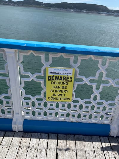 This outdoor shot shows a portion of the Llandudno Pier in Llandudno, United Kingdom. The camera angle is close, focusing on a railing and part of the pier's decking. A yellow warning sign is attached to the railing with the text: "Llandudno Pier BEWARE!! DECKING CAN BE SLIPPERY IN WET CONDITIONS". The pier railing has a geometric pattern in white with accents of blue. To the right, the wooden decking of the pier is visible. Beyond the railing, there is a view of the sea and a distant landmass. The image is slightly rotated clockwise.