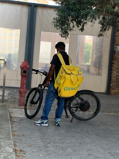 This is a vertical, eye-level shot of a person on a bicycle, possibly a delivery person. The individual is wearing a black t-shirt, blue jeans, and white and green athletic shoes. They have a large, bright yellow delivery bag strapped to their back, with the "Glovo" logo visible on it in green text. The bicycle is a dark color with black tires and appears to be an electric bike. In the foreground, a red fire hydrant is visible. The background consists of a building with large windows and brickwork above. The ground is paved with concrete and scattered with leaves. The scene appears to be outdoors, likely on a street or sidewalk. The lighting suggests it is daytime. The overall impression is a candid, everyday moment of a delivery person on duty.