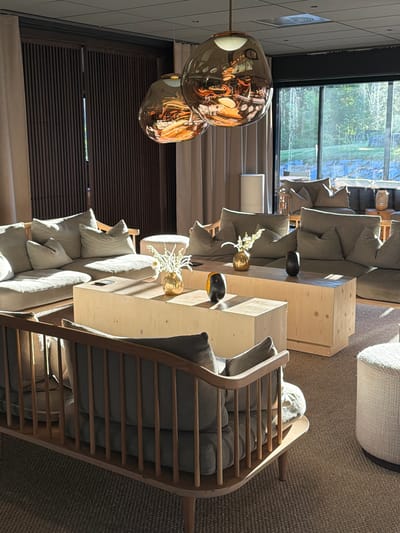 The image depicts the interior of a lounge area, likely within a hotel or resort, with a large window overlooking a natural, wooded landscape. The room features contemporary, comfortable seating arrangements, including several large, sectional sofas upholstered in a neutral fabric. Two wooden coffee tables, one significantly larger than the other and made of light-colored wood, are placed in front of the sofas. Decorative elements include a gold vase with white flowers on the larger coffee table, and a dark, oblong vase and a black and yellow decorative object on the smaller coffee table. Above the seating area, two large, irregularly shaped pendant lights with a metallic, reflective surface hang from the ceiling. The space is bathed in natural sunlight, casting strong shadows and highlighting the textures of the furnishings and the woven rug on the floor. In the background, a wooden slatted partition and sheer curtains add to the room's modern and sophisticated ambiance. The scene conveys a sense of relaxation and tranquility. The presence of contemporary furnishings and the panoramic window suggest it could be a high-end establishment. Given the query specifying Östersund, Sweden, this lounge area is situated in Östersund.