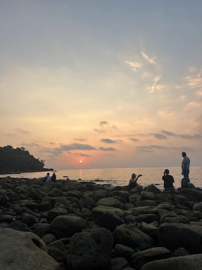 A panoramic view depicts a rocky beach in Preah Sihanouk, Cambodia, at sunset. The sky presents a gradient of light blue overhead, transitioning to orange and yellow hues near the horizon, featuring scattered stratocumulus clouds. The sun, a distinct orange disc, is positioned low above the ocean, partially obscured by clouds, casting a bright orange reflection path across the calm water. A verdant, tree-covered headland is visible on the left side of the frame, extending into the sea. The foreground is composed of numerous dark, irregular rocks, many displaying moss or lichen growth. Five individuals are situated on these rocks near the water's edge. On the left, two individuals are seated together, facing the sunset. On the right, one individual is seated, holding a smartphone horizontally towards the sunset, appearing to capture an image. Another person is seated adjacent to them, also facing the sea. To their right, a third person stands upright, observing the water, with a blue backpack resting on a nearby rock.