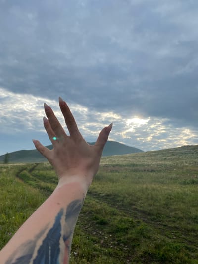 A close-up of a person's left hand and forearm is in the foreground, with the hand extended towards the sky. The hand has long, pointed fingernails and appears to have a faint green glint on the middle finger. The forearm is covered in a tattoo with blue and black ink. The background shows a grassy, hilly landscape with a winding dirt path. The sky is overcast with some patches of sunlight breaking through the clouds. The location is identified as Abzanovo, Russia.
