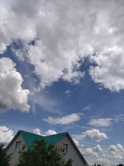 This is a vertical, eye-level, outdoor photograph of a house with a teal roof against a bright blue sky filled with large, fluffy white clouds and smaller wispy clouds. The house is light-colored, possibly white or pale grey, with a visible window on the left gable. Green foliage from trees partially obscures the lower portion of the house. The image appears to have been taken in the city of Nizhnyaya Makhtama, Russia, under a partly cloudy sky.