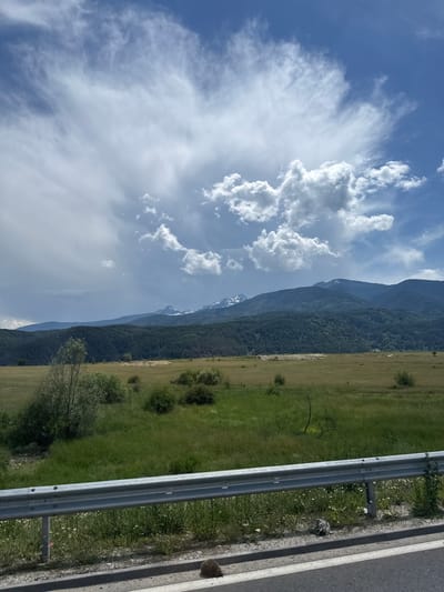 The image shows a landscape with mountains in the background and a grassy field in the foreground. A metal guardrail runs horizontally across the lower part of the image. A small hedgehog is visible on the side of the road, near the guardrail. The sky is partly cloudy with a mix of blue sky and white clouds. The location appears to be a rural area near Dobrinishte, Bulgaria.