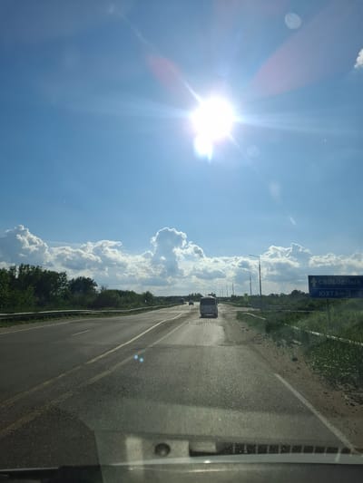 The image shows a view from a vehicle driving on a road. The road appears to be a highway with multiple lanes, indicated by the lane markings and guardrails. The sun is bright and high in the sky, creating lens flare effects and making it difficult to see details in some parts of the image. There are several cars and a bus on the road ahead. On the right side of the road, there is a blue sign with white text in Russian, which appears to be a road sign indicating directions and distances to towns. The text on the sign reads "СВОБОДНЫЙ" (Svobodny) and "ЮХТА - 3 0" (Yukhta - 3 0). This suggests the location is near the town of Yukhta and possibly heading towards Svobodny. There are trees and vegetation along the sides of the road, and fluffy white clouds are visible in the bright blue sky. The overall environment appears to be rural or semi-rural, consistent with a highway outside of a major city. There is no indication of any people outside of the vehicles.