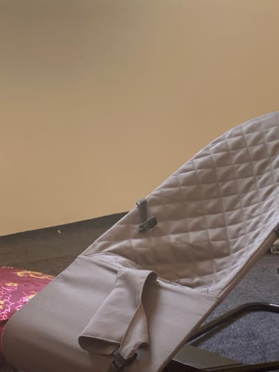 The image shows a close-up of a beige fabric baby bouncer. The bouncer has a quilted pattern on the seat and a folded fabric piece near the bottom. A metal frame is visible beneath the fabric. The background consists of a wall painted a light tan color and a dark carpeted floor. To the left of the bouncer, a portion of a pink and gold patterned textile is visible, possibly a blanket or cushion. The scene is indoors and appears to be a domestic setting. The image does not contain any information that would allow for the determination of its location within the city of Arbagish, Uzbekistan.