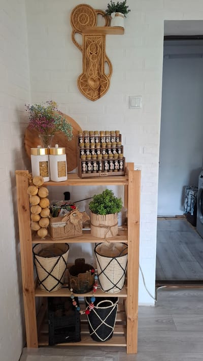 This is a full-shot, eye-level, indoor photograph of a wooden shelving unit filled with various decorative and practical items. The shelving unit is placed in a corner with a white brick-patterned wall behind it.
On the top shelf, a wooden wall decor piece with intricate carvings is mounted, featuring a heart shape at the bottom. A small white pot with a sprig of lavender sits on a small shelf attached to the decor. Below this, on the top shelf of the unit, are two tall, white jars with gold lids containing pasta, and a tiered wooden spice rack filled with numerous small spice jars. To the left of the jars, a vase of dried, multi-colored baby's breath flowers is visible.
The second shelf down holds a string of garlic bulbs hanging on the left. In the center, there's a small woven basket with a burlap flower decoration and another decorative element with a tag. To the right, a potted green plant in a wooden cylinder-like container is present, tied with twine.
The third shelf from the top contains two woven baskets with black criss-cross patterns. Between them sits a dark, rustic-looking pot with a colorful beaded garland draped around it.
The bottom shelf has a black plastic crate filled with items, possibly potatoes or other produce. To the right of the crate, another woven basket with a similar black criss-cross pattern is present, this one being black with white accents.
The floor appears to be light gray laminate flooring. To the right of the shelving unit, a doorway leads to another room, where a washing machine and some laundry are visible. The overall impression is one of a cozy and organized kitchen or pantry area.
The image is described as being from Cherkessk, Russia. However, there are no visual elements in the image that specifically identify it as being in Cherkessk, Russia. The description is based solely on the visual content of the image.