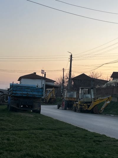 This image depicts a rural or semi-rural street scene at dusk or dawn in Bukovlak, Bulgaria. The sky transitions from a pale blue overhead to an orange and pink hue near the horizon. No individuals are present.
In the foreground, a patch of green grass occupies the lower left. A weathered, light blue dump truck is parked partially on the grass and partially on the paved road, its rear facing the viewer. Its tailgate is closed.
Mid-ground, a grey asphalt road extends diagonally from the lower right towards the center. Parked along the right side of this road are two yellow heavy equipment vehicles. The foremost is a yellow tractor with a visible red rear wheel. Immediately behind it is a yellow backhoe loader with its rear digging arm extended downwards, the bucket resting near the ground. The brand "TERRA" is visible on the backhoe arm. Further along the road, a third yellow backhoe loader is partially visible near a house on the left.
In the background, a white house with a brown tiled roof and a chimney with a metal structure is visible on the left. Another house with a darker tiled roof is present on the right. Multiple utility poles, including one with a street light fixture on the right, support numerous overhead power lines that crisscross the sky. Deciduous trees, currently bare, are visible around the houses.