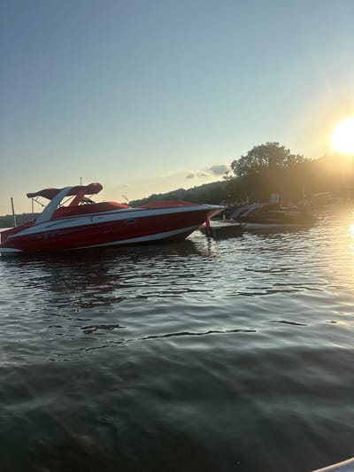 In Algonquin, United States of America, a red and white Crownline boat is docked on a body of water, likely a lake or river, under a clear sky with a warm sun setting in the background. The boat is covered by a red canopy. In the background, there are other boats docked, including jet skis, and a tree-lined shore. The water is calm with gentle ripples, reflecting the sunlight.