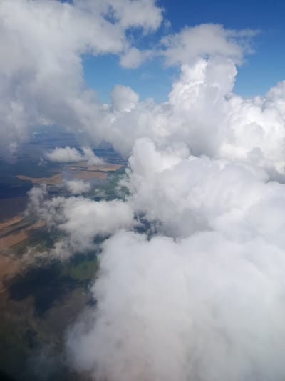 An aerial photograph captures a landscape dotted with agricultural fields and patches of vegetation, partially obscured by a layer of cumulus clouds. The sky above is a clear blue, with pockets of white clouds scattered across it. The ground below reveals a mosaic of agricultural land, with fields of varying shades of brown and green, suggesting different crops or stages of growth. Some areas appear to be recently plowed, while others show established vegetation. Shadows cast by the clouds create a dappled effect on the landscape, adding depth and dimension to the scene. The photograph appears to be taken from a high vantage point, possibly an aircraft, given the perspective and the vastness of the landscape visible. The location is identified as Teplovka, Russia.