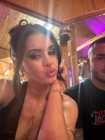 This image captures an indoor scene, likely a restaurant or bar, located in Debrashtitsa, Bulgaria. The foreground is dominated by a young woman posing for the camera, occupying the left and center of the frame. She has dark hair styled in a messy bun with a distinct loop on top and some loose strands framing her face. Her eyes are heavily made up with dark shadow and liner, and her eyebrows are well-defined. She is making a puckered, pouting expression with full lips, looking directly into the camera. She wears gold-toned hoop earrings, a delicate gold chain necklace with a small, elongated pendant (resembling a key or bar), a thick gold-toned chain-link bracelet on her right wrist, and a thin gold bangle on her upper right arm. Her right hand is raised, with fingers gently touching her neck/chin area. She appears to be wearing a dark, possibly brown or black, sleeveless or halter-style top.
To her right, partially visible, is a young man. Only the right side of his face and upper torso are visible. He has short, dark hair with a faded cut on the sides and a short, dark beard. He is wearing a black t-shirt with a design on the chest featuring red and blue lettering, part of which reads "DID". His gaze is directed slightly downwards and away from the camera.
The background features light-toned wooden paneling on the walls and a sloping wooden ceiling. Various light sources illuminate the scene: a prominent bright light flare (possibly from a window or strong artificial light) is visible at the top center, casting a strong glare. On the left side of the background, a horizontal artificial light source is visible. On the far right, parts of the wooden ceiling structure are illuminated with a distinct purple/magenta light. A small, decorative wire cage with artificial greenery and flowers is suspended from the wooden ceiling towards the upper left background. Further in the background, behind the man, another individual wearing a light-colored t-shirt and trousers is faintly visible, standing.