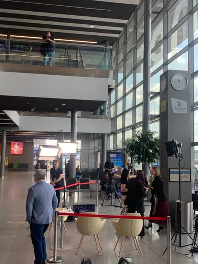 The image shows the interior of Katowice Airport in Pyrzowice, Poland (latitude: 50.471, longitude: 19.072). The scene is set up for a television interview. A man in a dark suit is standing in the foreground, facing a camera. Behind him are two white chairs and a laptop computer set up on a small table. Three women are seated on the chairs in front of the man, and one of them is speaking to the camera. Behind them are additional camera and lighting equipment, a television screen, and a large potted plant. In the background, a man in a blue shirt is standing behind a table while a woman in a black dress and high heels speaks on her cell phone. Two men are seated in the background, watching the interview. The scene is illuminated by overhead lights and studio lighting equipment. The airport terminal is modern and clean with large windows and a high ceiling. The camera is positioned at a medium distance and at eye level. The image is clear and well-lit.