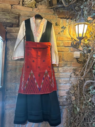A traditional Bulgarian folk costume is displayed against a brick wall, adorned with a street lamp and climbing ivy. The costume consists of a white shirt with embroidered trim, a green vest, a red woven apron with geometric patterns, and a black skirt. The location is in the city of Kalimantsi, Bulgaria.