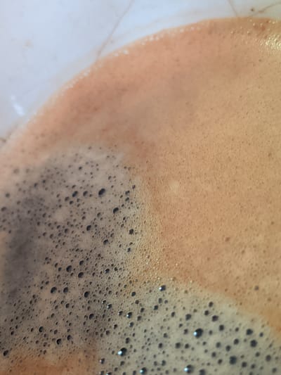 The image is a close-up, overhead shot of the foamy surface of a dark liquid, likely coffee, filling a portion of a light-colored cup. The foam is varied, with larger, darker bubbles in the lower left transitioning to a smoother, lighter tan foam in the upper right. There is no indication of the city of Galiche, Bulgaria, or any other specific location.