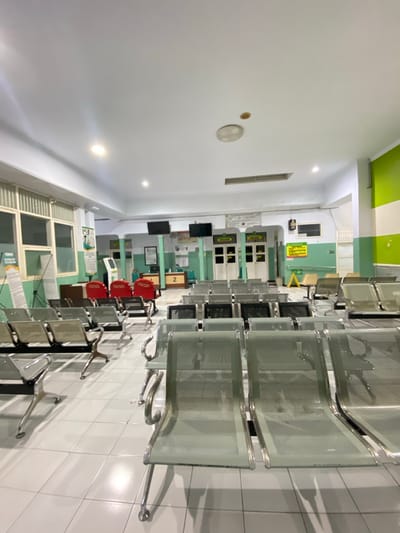 The image depicts a waiting area with rows of empty metal chairs arranged on a tiled floor. The walls are painted in a combination of white and light green. In the background, there are windows with vertical blinds, a reception desk with a sign showing the number "2", and several doorways. There are also two large screens mounted on the wall. The lighting appears to be artificial, with recessed lights in the ceiling. The scene suggests a public space, possibly a clinic or a government office, located in Margoyoso, Indonesia.