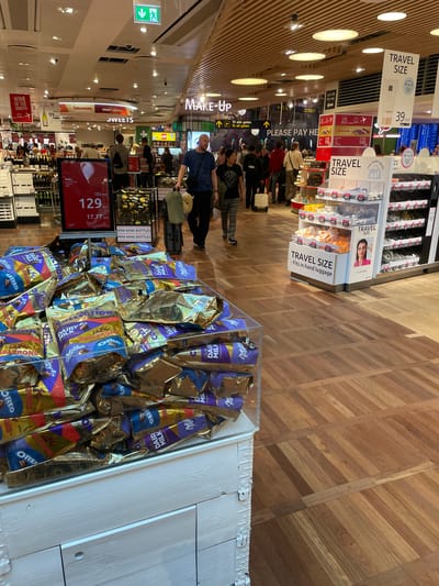 The image depicts the interior of a duty-free shop at Copenhagen Airport (CPH) in Tårnby, Denmark. A prominent display of chocolate bars, including Milka, Toblerone, and Oreo-branded products, dominates the foreground. A digital display shows a price of 129 DKK (17.77 EUR). A queue of customers is visible near the checkout area, indicated by "PLEASE PAY HERE" signage. The shop features products categorized as "SWEETS", "MAKE-UP", "WINE OF THE MONTH", and "TRAVEL SIZE". The floor is made of light-colored wooden planks. The general atmosphere indicates a busy retail environment within an airport terminal.