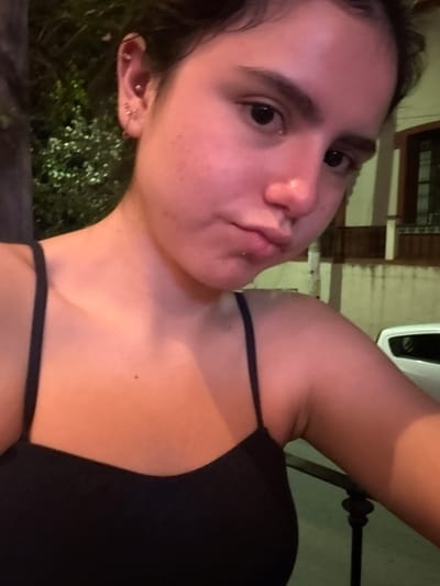 A young female individual is depicted in a close-up, waist-up portrait, looking directly at the camera. Her dark hair is pulled back, revealing multiple piercings on her left ear, including a helix piercing and several lobe piercings with small rings. Her dark eyes are open and focused forward. Her skin exhibits minor blemishes on the cheeks and chin, and a small white substance is visible on or below her lower lip. She is wearing a dark, spaghetti-strap top, with a visible tan line on her left shoulder. The scene is outdoors at night or in low-light conditions, with artificial lighting casting an orange hue on her skin. The background to the left features blurred dark foliage. To the right, a building with a light-colored wall and a dark-framed window is visible, along with a dark railing. A white vehicle is partially discernible behind the railing. The location is Santiago, Mexico.