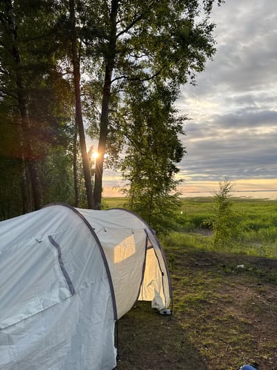 A white tent is pitched on a patch of grass and dirt, with its entrance open and illuminated from within. Trees with green foliage are visible in the foreground and on the left, with sunlight filtering through them. In the background, a body of water stretches to the horizon under a cloudy sky. The scene appears to be a campsite, possibly located in Bolshaya Izhora, Russia, given the natural landscape and proximity to water. There are no people visible in the image.