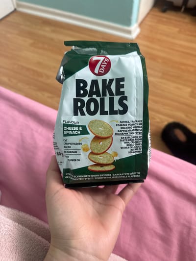 A person is holding a bag of 7 Days Bake Rolls, cheese and spinach flavor. The bag is white with green accents and features an image of the bake rolls. The person's hand is visible, and a pink fabric surface is in the background. The background also shows part of a wooden floor. The image does not show the city of Armagh, United Kingdom.