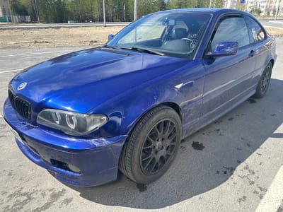 A blue two-door BMW car, likely a 3-series (E46 coupe), is parked on an asphalt surface under bright daylight. The car appears dirty, covered in dust and grime, particularly on the hood, front bumper, side panels, and exterior mirrors. The front bumper also shows scuffs and minor damage. Its headlights are slightly cloudy. The car features black multi-spoke wheels with "MOTORSPORT" text visible on the hub caps. A white sticker is adhered to the rear passenger-side window.
The scene depicts a semi-urban or fringe area. In the background, there is a line of coniferous trees, possibly pines, and an unpaved or construction-like area with some dirt. Utility poles or streetlights are visible among the trees. Further in the distance, apartment buildings can be seen, suggesting a town or city. The asphalt ground beneath and around the car has various dark stains and dirt. Strong shadows indicate a sunny day, likely mid-morning or early afternoon. The location is specified as Gubkinsky, Russia.