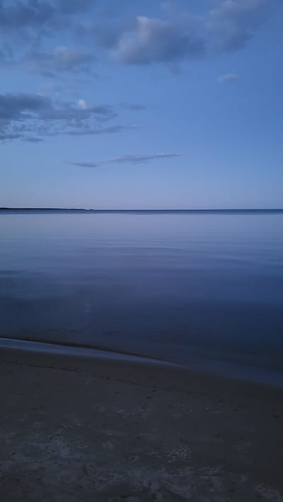 This is an outdoor, vertical, eye-level, medium shot of a twilight scene at the beach in Zaostrov'e, Russia. The sky is a deep, clear blue with scattered, wispy clouds. A thin, dark horizon line separates the sky from the calm, vast expanse of water. The water is a muted blue, reflecting the dim light of dusk. The foreground features a sandy beach with subtle indentations, suggesting footprints or the texture of the sand. The scene evokes a sense of tranquility and solitude.