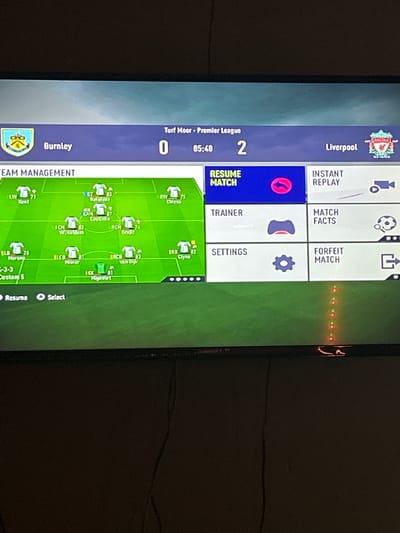The image is a photograph of a television screen displaying a menu from the FIFA video game. The game shows a match between Burnley and Liverpool in the Premier League, with Liverpool leading 2-0 in the 85th minute. The menu includes options for resuming the match, viewing an instant replay, accessing the trainer, viewing match facts, adjusting settings, and forfeiting the match. Also visible on the screen is a team management display, showing the player formations for Liverpool. There are no people visible in the image, only the television screen and a dark background. There is no discernible connection to the city of Kalitinovo, Bulgaria.