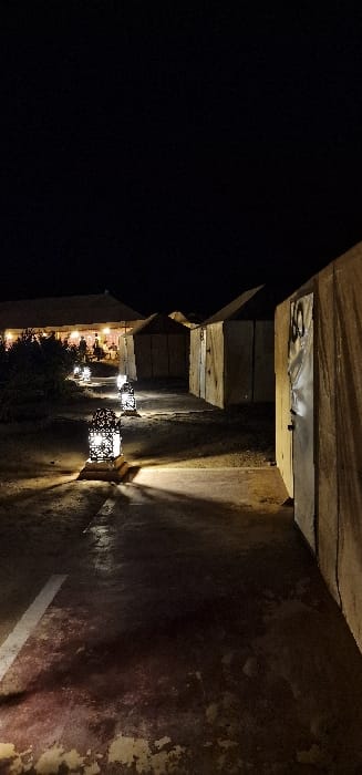 The image displays a nocturnal scene at a desert camp in Et-taous ⵟⴰⵡⵙ الطاوس, Morocco. A reddish-brown pathway with a partial white stripe on the left foreground extends into the background, illuminated by a series of five ornate, perforated metal lanterns placed at intervals along its left edge. These lanterns cast distinctive light patterns and shadows on the ground.
On the right side of the pathway, a row of light-colored, canvas-like tent structures, presumed to be accommodation units, is visible. The closest tent on the right features a partially obscured number, possibly "10" or "60," on its entry flap. To the left of the pathway, dark, low-lying vegetation is present. In the distant background, a larger, illuminated tent structure is visible, within which several indistinct human silhouettes can be observed, indicating the presence of people engaged in activities. The sky is uniformly dark, consistent with a nighttime environment. No additional objects, specific actions, or direct interactions are clearly discernible.