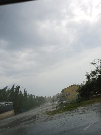The image captures a stormy day in the city of Kumush-Aziz, Kyrgyzstan. The sky is overcast with heavy, grey clouds, and streaks of rain are visible, indicating a downpour. The scene depicts a road with a gravelly surface, appearing wet and reflective. On the left side of the road, a low-rise building with a dark facade and a lighter-colored base is visible. Further down the road, a line of tall, thin trees with wind-swept foliage borders the landscape.

A yellow, multi-story building stands on the right side of the road, with several windows visible. Adjacent to the yellow building, dense green trees and foliage create a natural backdrop. The road itself appears to be a main thoroughfare, with a red car visible in the distance, driving away from the viewer. The overall impression is one of a rural or semi-urban area experiencing inclement weather. The composition suggests the photo was taken from a vehicle, looking out through a rain-streaked window.