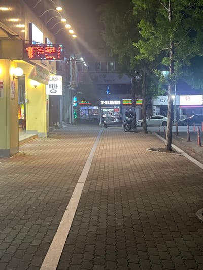 The image captures a night scene on a city street in Gwangju, South Korea. A paved pedestrian walkway, made of interlocking dark gray bricks with a white line marking its edge, stretches into the distance. Buildings line the left side of the walkway, displaying illuminated signs, including a red LED sign with Korean characters. A yellow-painted storefront is prominent on the left. Across the street, trees with lush green foliage stand tall, their branches reaching towards the night sky. In the background, more buildings and businesses are visible, including a 7-ELEVEN store with its distinctive branding. Vehicles, including a white car and a motorcycle, are parked or stopped along the street. The overall impression is that of a typical urban commercial area after dark, illuminated by artificial lights from businesses and streetlights.
