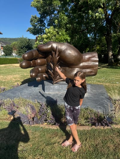 In Divonne-les-Bains, France, a young girl stands in front of a large bronze sculpture of a hand holding a wheat stalk. She is wearing a black t-shirt and patterned shorts, with pink slides on her feet. The sculpture is placed on a dark grey, rectangular base in a grassy area with some low-lying plants. Behind the sculpture, trees and a blue sky are visible, and in the distance, buildings and a hillside can be seen. The sun casts shadows on the ground.