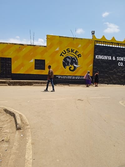 The scene depicts a daytime street view in Nkuene ward, Kenya, under a clear blue sky with sparse white clouds. In the background, a building with a prominent yellow facade features diagonal yellow stripes and a black base. A large black "TUSKER" logo, including an elephant silhouette, is centered on the yellow wall. Below the logo, white text warns, "EXCESSIVE ALCOHOL CONSUMPTION IS HARMFUL TO YOUR HEALTH. NOT FOR SALE TO PERSONS UNDER THE AGE OF 18 YEARS." To the left of the logo, a black rectangular door is visible, and a small black window is to its right. Adjacent to this, a black wall with yellow crenellated decorative elements along its roofline displays "KINGINYA & SON'S CO." in white text.
In the foreground, a light brown paved road occupies the majority of the frame. A concrete curb, soiled and showing some dry debris, runs along the left edge of the road. Three individuals are present:
1. A male subject, positioned left of center, wears a brown jacket, ripped blue jeans, and dark sneakers. He is walking from left to right across the road.
2. A female subject, located further right and closer to the building wall, wears a purple skirt or dress and a lighter top. She is walking from left to right along the edge of the road, parallel to the building.
3. A second female subject is partially visible further to the right and slightly behind the second individual, wearing dark clothing and brown shoes, also walking from left to right along the building wall.
No direct interactions are observed between the individuals. The overall environment appears to be a commercial or residential street.