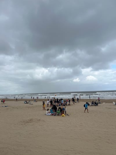 The image captures a crowded beach scene under an overcast sky in Punta del Este, Uruguay. The broad expanse of light brown sand stretches across the lower half and right side of the frame, meeting the ocean with moderate waves breaking into white foam along the waterline. The sky, dominating the upper left and central parts of the image, is filled with dense, dark gray clouds, suggesting a cloudy or potentially stormy day, though daylight is clearly present.
Numerous people are enjoying the beach. Many are gathered in groups, seated on various colorful beach chairs, some accompanied by partially closed beach umbrellas. These groups appear to be relaxing, conversing, or simply observing the ocean. One prominent figure in the foreground, wearing a blue top and dark shorts, walks along the sand, seemingly heading towards the water. Closer to the ocean's edge, more individuals are visible, some standing in the shallow water, others scattered along the sand. A surfboard is propped upright in the sand near the water, indicating watersports activity. A small, orange marker or flag is also visible near the waterline. Despite the cloudy weather, the beach is lively, reflecting a relaxed atmosphere of people enjoying the coastal environment. No visible text is discernible in the image.