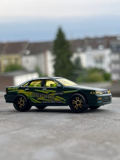 This is a close-up, side-view, rotated image of a green toy car, possibly a model of a Nissan Cefiro, resting on a textured, grey surface. The car has yellow-tinted windows and gold-colored wheels with a star-like spoke pattern. A stylized yellow and green design adorns the car's side, with the text "CEFIRO" prominently displayed. Below "CEFIRO," the website "WWW.MAJORETTE.COM" is visible, and further down, the text "BILSTEIN" is present. The background is blurred, showing residential buildings and greenery, suggesting an outdoor setting. The lighting appears to be natural daylight. The image is taken in Neuss, Germany.