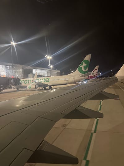 The image displays a nighttime scene at an airport, viewed from inside an aircraft. A large wing dominates the right side of the frame. Beyond the wing, another plane, identifiable by its "transavia" logo and green tail, is parked near a terminal building with bright lights. A Turkish Airlines aircraft's tail is visible further in the background. The area appears well-lit despite the darkness.