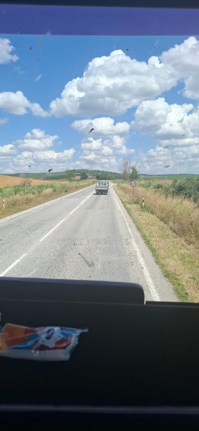 The image captures a view from inside a vehicle looking out through a windshield. The windshield is dirty with numerous small dark specks, likely insects. In the distance, a paved road stretches forward through a rural landscape. The road has white dividing lines and appears to be in fair condition with some patches. A light-colored pickup truck is driving on the road ahead, moving away from the camera. To the right of the road, a circular traffic sign with red and white markings is visible, indicating a restriction. The surrounding environment consists of dry, grassy fields and some scattered trees and vegetation, with rolling hills in the background under a bright blue sky filled with fluffy white clouds. The lower part of the image shows the interior of the vehicle, including a dark dashboard and a partially visible package with colorful wrapping. The scene appears to be on a sunny day in the countryside. The location is identified as being on a road near the city of Karaisen, Bulgaria.