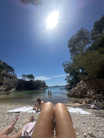 The image presents a first-person perspective scene at Cala Fornells beach in Peguera, Spain. The viewer is situated on a pebble beach with a partial view of their own legs, and partially other person's feet. In the background, the turquoise water of the cove is visible, with several people swimming or wading. The beach is bordered by rocky cliffs covered with trees and vegetation. The sky is clear and bright blue, with the sun shining intensely, creating glare. One can see towels, bottles, sunglasses and other personal items on the beach nearby.