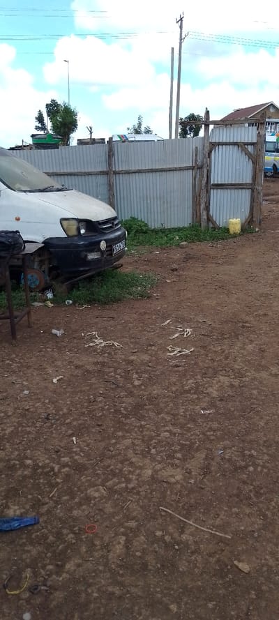 The scene is an unpaved outdoor area in Kuku ward, Kenya, under a partially cloudy sky. The ground consists of reddish-brown dirt mixed with small stones and scattered debris, including white plastic strips, a blue plastic bottle, a red rubber band, and a yellow wire.
In the midground, the front portion of a white van with a black bumper is visible on the left. The vehicle's front passenger-side headlight assembly is absent, revealing mechanical components, and the front passenger-side wheel is missing, exposing a blue brake rotor/drum. The license plate "KBAL 152" is affixed to the front. Green vegetation surrounds the vehicle's base. A dark bag rests on a metal frame to the left of the van.
A grey corrugated galvanized iron fence with wooden posts and crossbeams extends across the midground from behind the van to the right. A wooden gate is integrated into the fence on the right. A yellow plastic jerrycan is located on the ground adjacent to the fence.
In the background, a building with a reddish-brown sloped roof is partially visible to the right. A white public transport minibus (matatu) with red, green, and blue stripes and "BESTCOACH" written on its side is parked behind the fence. Multiple utility poles with overhead power lines are present, and a street light pole stands in the upper left background.