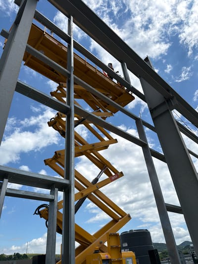 A low-angle, outdoor shot captures a construction site with a yellow scissor lift extending upwards amongst unfinished steel beams. A worker in a hard hat and safety vest is visible in the platform of the scissor lift, appearing to be engaged in work. The sky above is blue with scattered white clouds. In the background, rolling green hills and trees are visible, with a cell tower in the distance. A large black water tank with "PRO" written on it sits near the base of the scissor lift. The scene suggests ongoing construction activity in a semi-rural or suburban area.