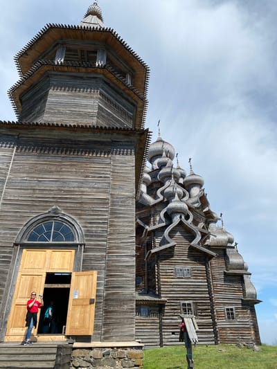 This is a low-angle, exterior shot of a large, intricate wooden church, likely the Transfiguration Church on Kizhi Island, in Patanevshchina, Russia. The church is constructed entirely of wood, with a tall, multi-tiered bell tower on the left and a series of onion domes on the right, characteristic of Russian Orthodox architecture. The wood appears weathered and aged. A woman in a red shirt and jeans stands on the steps to the left, holding a phone to take a picture. She is partially obscured by the open wooden doors. Another person, wearing a teal top, is visible inside the doorway. The church is situated on a grassy hillside under a cloudy sky.