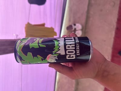 A hand holds a black energy drink can with green and purple jagged patterns. The can has the brand name "GORILLA ENERGY DRINK" written in white text. A circular sticker with a QR code and some text is affixed to the can. The background is blurred and appears to be an indoor setting with a pinkish-purple light source. There are some small pink and white objects in the background, possibly shoes.