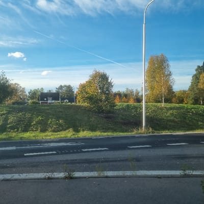 This image depicts a daytime roadside scene in Västervik, Sweden, during an autumn period.
The lower third of the frame shows an asphalt roadway with broken white lane markings and a continuous white line delineating a paved shoulder or curb. Sparse vegetation, primarily small weeds, is visible along the pavement edge. Rising from the roadway is a grassy embankment that occupies the middle ground. A white lamppost, featuring a curved upper arm, is positioned on the right side of the embankment, emerging from the grass.
Numerous deciduous trees are present on and beyond the embankment, displaying autumnal foliage in shades of green, yellow, and orange. A prominent tree with yellow-orange leaves stands in the central-right. In the background, positioned on the crest of the embankment in the left-center, is a two-story house with red siding, a dark gabled roof, and white window trim. A flagpole is visible near the house. Additional trees form a denser treeline behind the house and extend across the background.
The upper portion of the image reveals a clear blue sky with scattered wispy white clouds and a distinct, straight white contrail traversing diagonally from the upper left to the mid-right. No individuals or specific actions are depicted within the scene.