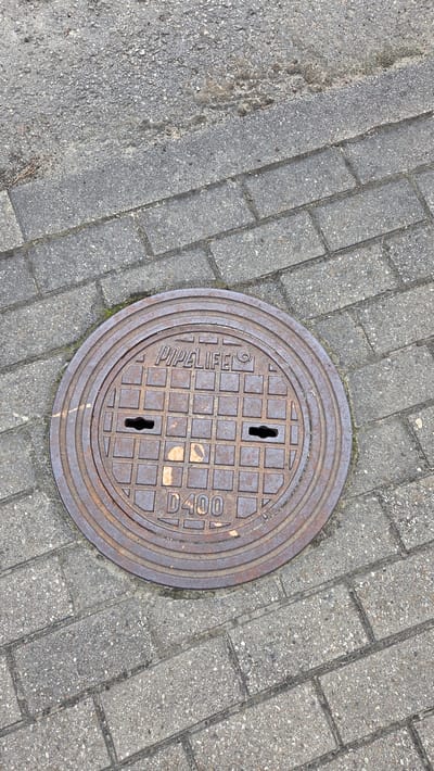This is a top-down, close-up, eye-level shot of a manhole cover set into a paved surface. The manhole cover is round and made of dark, rusted metal with concentric rings around its edge. The surface of the cover features a grid pattern of raised squares, and two keyhole-shaped openings are visible. The text "PIPELIFE" is embossed in capital letters on the left side of the manhole cover, and "D400" is embossed on the right. The surrounding ground is paved with rectangular, grey interlocking pavers. To the left of the pavers, there is a strip of rougher, darker asphalt or concrete, with some visible debris and dried vegetation. The lighting suggests it is daytime. No people are visible in the frame. The scene appears to be an outdoor urban or suburban setting, likely a street or sidewalk in Ūlupji, Latvia.