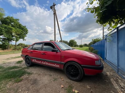 A red Volkswagen Passat sedan is parked on a dirt road next to a blue corrugated metal fence and gate. The car has a sticker on its side that reads "Passat club krasnodar." A utility pole with a lamp is visible behind the car, with power lines extending across the frame. Trees and green foliage are present on the left side of the image, and a larger tree with dense leaves overhangs the top right corner. The sky is cloudy with patches of blue. The scene appears to be in a rural or semi-rural setting, possibly the village of Otrado-Olginskoye, Russia, given the context of the sticker.