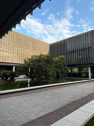 This vertically oriented image captures a modern architectural landscape under a bright, sunny sky with scattered white cumulus clouds, in what is identified as Preara, Italy. The scene features two distinctive buildings, a reflective water body with lush greenery, and geometrically arranged ground surfaces.

To the left, a tall building presents a facade of numerous horizontal metallic or grey slats, possibly sun shades or louvers, creating a textured pattern. Adjacent to it, another building stands with a warm, light brown or beige facade, composed of vertical textured panels.

The central part of the image is dominated by a long, narrow water feature with green-tinged water, reflecting the surrounding structures and sky. Several green trees and various types of foliage, including a prominent dense-leafed tree and some palm-like plants, grow along the edge of this water. White, cylindrical support pillars are visible, appearing to rise from or beside the water, adding to the structural aesthetic.

On the right, a wide paved area features a pattern of small, grey, square or diamond-shaped cobblestones. This paving is bordered by a strip of dark, granular gravel, which in turn lies next to another section of lighter grey, possibly textured concrete or stone.

In the bottom left corner, a dark, angular architectural element, possibly an overhang or part of a roof structure, extends into the frame, characterized by a series of sharp, triangular projections along its edge.

The overall impression is one of a meticulously designed and well-maintained outdoor space, perhaps a courtyard or public area within a modern complex. The time of day is clearly daylight, indicated by the bright illumination and visible shadows. There are no people or visible text in the image.